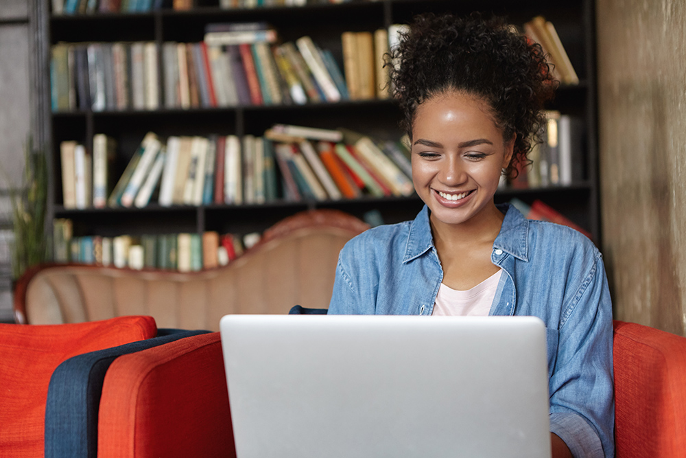 People, technology and education concept. Happy pretty dark-skinned female in jean shirt sitting at sofa using laptop, typing something and browsing internet while being in reading hall or library.